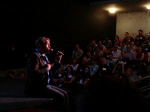Sex and relationship expert Reid Mihalko of ReidAboutSex.com sitting on a dark stage in a dark suit and blue shirt speaking to a full house at a MindshareLA event